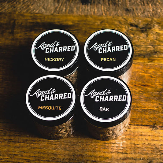Four containers labeled Aged & Charred with different types of wood chips: pecan, mesquite, hickory, and oak, arranged on a wooden surface.