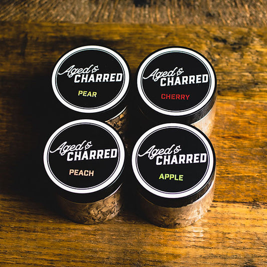 Four containers labeled Aged & Charred with different wood types: apple, cherry, pear, and peach, arranged on a wooden surface.