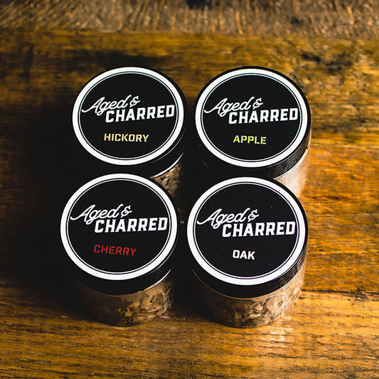 Four containers labeled Aged's Charred with different types of wood chips including Hickory, Apple, Cherry, and Oak, arranged on a wooden surface.